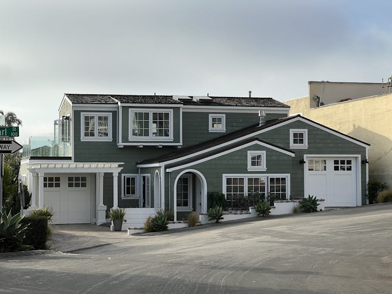 A stylish green two-story suburban house with white trim and tilt-up garages on a sunny street corner.