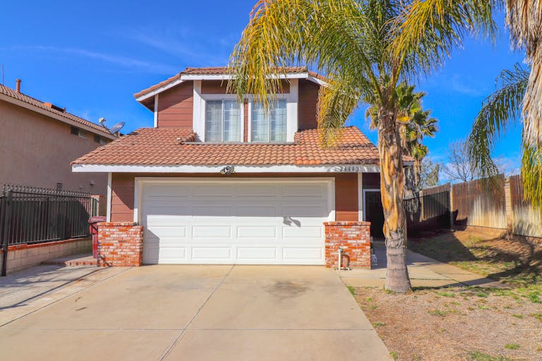 A sunlit suburban house with a garage and palm trees in the driveway during the summer heat.