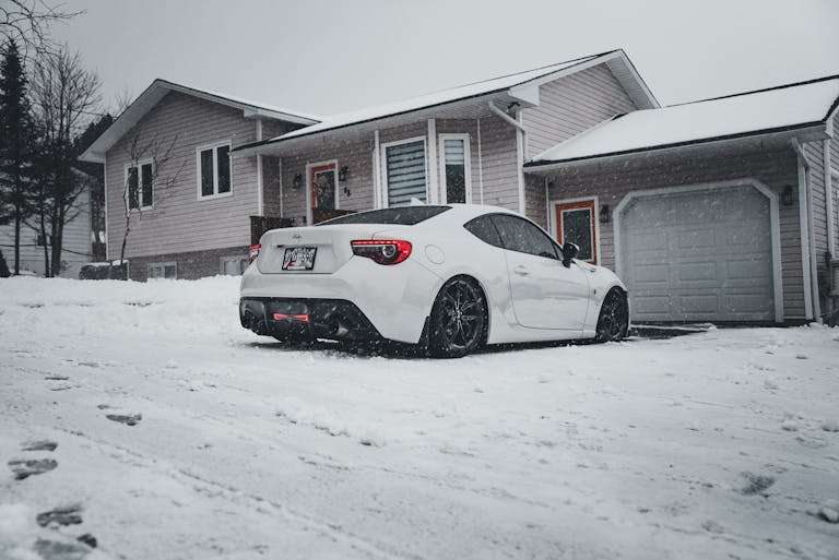 A white car parked in front of garage during winter snowfall.