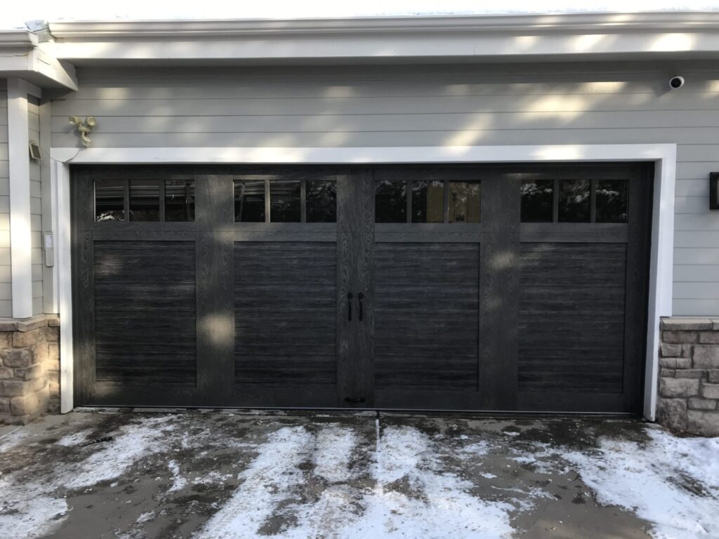 Black 4 panel custom garage door featuring split windows, faux barn style on a new home in the winter months, snow covered driveway.